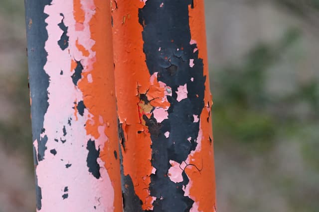 Close-up of weathered orange and black paint peeling off a metal pole with a blurred background