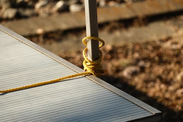 Close-up of a dock secured with yellow cord tied around a metal pole, with blurred gravel ground in the background