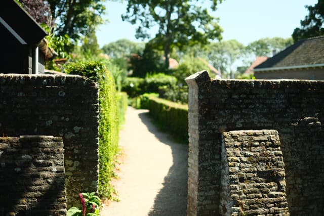 Narrow dirt pathway bordered by brick walls and lush green hedges, leading toward distant trees under bright daylight