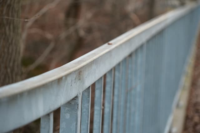 Close-up of a faded blue metal railing running along an outdoor path with a soft, blurred background of trees