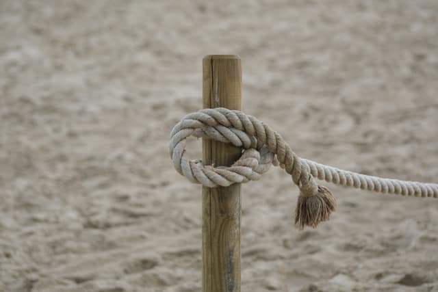 Close-up of a knotted rope tied around a wooden post on sandy ground