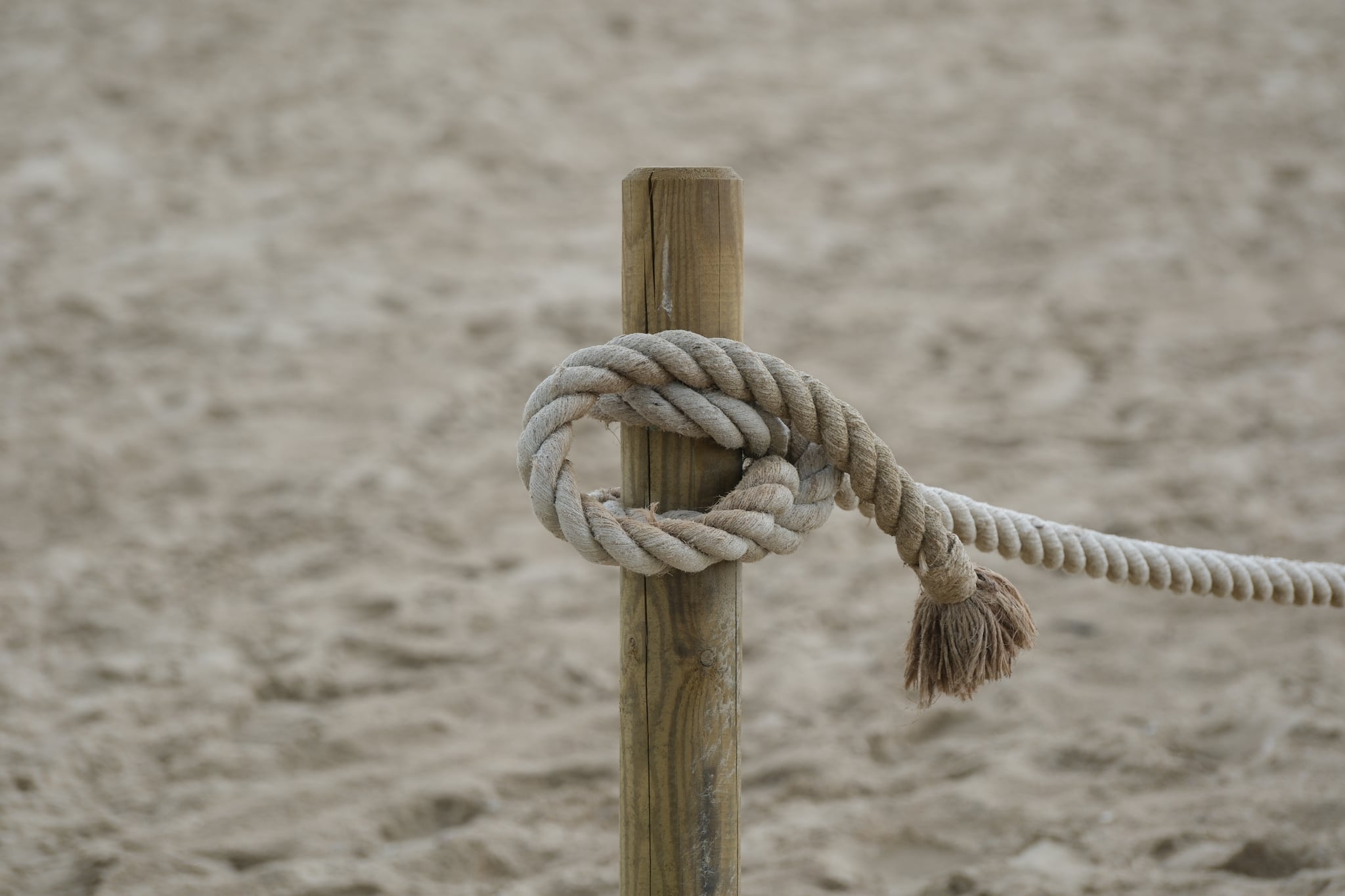 Close-up of a knotted rope tied around a wooden post on sandy ground